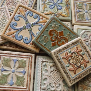 Può includere: Una collezione di piastrelle quadrate in ceramica con vari motivi in rilievo nei toni del blu, verde, marrone e crema. Ogni piastrella presenta intricati disegni a volute, motivi floreali e bordi geometrici con una finitura lucida e multicolore.