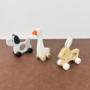 Op de afbeelding: Drie houten dierenspeeltjes: een witte hond met een zwart oor, een witte gans met een oranje snavel en een bruin konijn. Elk speelgoed heeft wielen en een uitgesneden ontwerp. Het speelgoed staat op een bruin oppervlak.