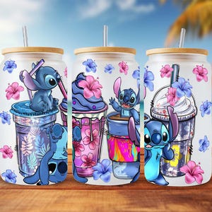 May include: Three glass tumblers with bamboo lids and straws, each featuring a cartoon Stitch design with tropical flowers. The tumblers have a frosted appearance and are decorated with blue and pink floral accents, perfect for cold drinks.