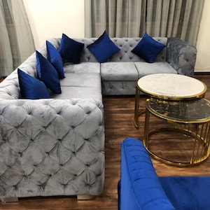 May include: A grey, button-backed corner sofa with blue velvet cushions. Two round side tables with gold frames and marble tops are in front of the sofa. A blue velvet armchair is in the foreground.
