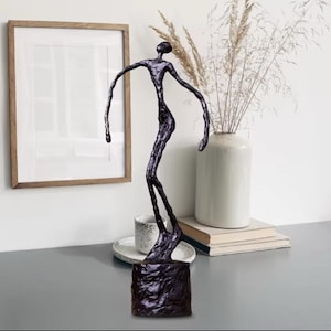 May include: A dark bronze sculpture of a human figure with outstretched arms. The abstract art piece is mounted on a textured, square base. The sculpture is displayed on a grey surface with a framed picture, vase, and books in the background.