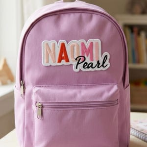 May include: A light purple backpack featuring a large, multi-colored fabric patch on the front. The patch displays the name NAOMI in block letters with a red A, and the word Pearl in black cursive script. The bag has a front zippered pocket.