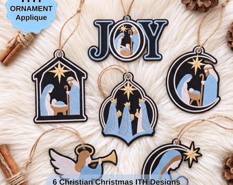 May include: Six embroidered Christmas ornaments featuring nativity scenes, the word JOY, and an angel with a trumpet. Each ornament has a black background with light blue and gold accents, a hanging twine loop, and intricate stitched details.