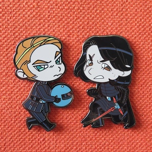 May include: Two enamel pins featuring characters from Star Wars. The pin on the left is a light-skinned character with brown hair and a blue and white sphere. The pin on the right is a dark-skinned character with black hair and a red lightsaber.