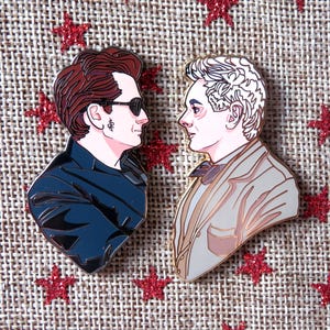 May include: Two enamel pins showing side profiles of characters. One pin features a person with sunglasses and a black jacket, the other a person in a suit. Both pins have gold outlines and are set against a textured background with red star accents.