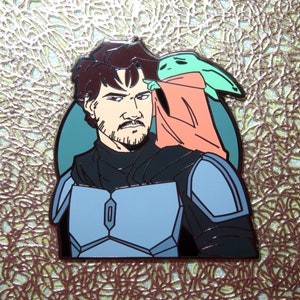 May include: A black and blue enamel pin featuring a man with brown hair and a beard, wearing a black and blue suit, with a green and pink alien character on his shoulder.