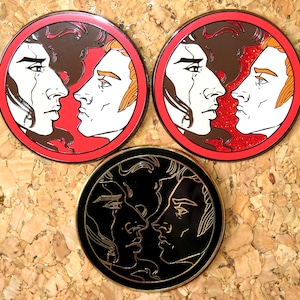 May include: Three round pins with black outlines and different colored backgrounds. The first pin has a red background with two men's faces looking at each other. The second pin has a red background with two men's faces looking at each other. The third pin has a black background with two men's faces looking at each other.