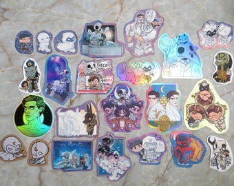 Peut inclure: Une collection d'autocollants découpés assortis représentant divers personnages illustrés, incluant des figures en costumes blancs à capuche, des divinités à tête d'animal et des portraits style chibi aux designs graphiques holographiques.