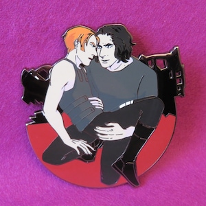 May include: A red, white, and black enamel pin featuring two men embracing. The man on the left has orange hair and is wearing a black vest. The man on the right has black hair and is wearing a black shirt. The pin is set against a red background with a black silhouette of a building in the background.
