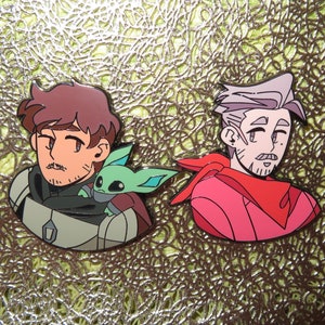 以下が含まれることがあります： 漫画のイラストが描かれた2つのエナメルピン。1つのピンは、茶色の髪の男性と小さな緑色の生き物を描いています。もう1つのピンは、灰色の髪と赤いスカーフの男性を示しています。ピンは、テクスチャ加工された金色の表面にあります。