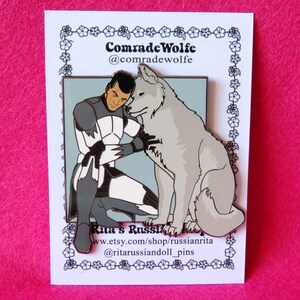 May include: A white enamel pin featuring a cartoon illustration of a man in a gray and white uniform with a white wolf. The man is kneeling and hugging the wolf. The pin has a blue background and the text "Comrade Wolfe @comradewolfe" at the top and "Rita's Russian Army www.etsy.com/shop/russianrita @ritarussiandoll_pins" at the bottom.