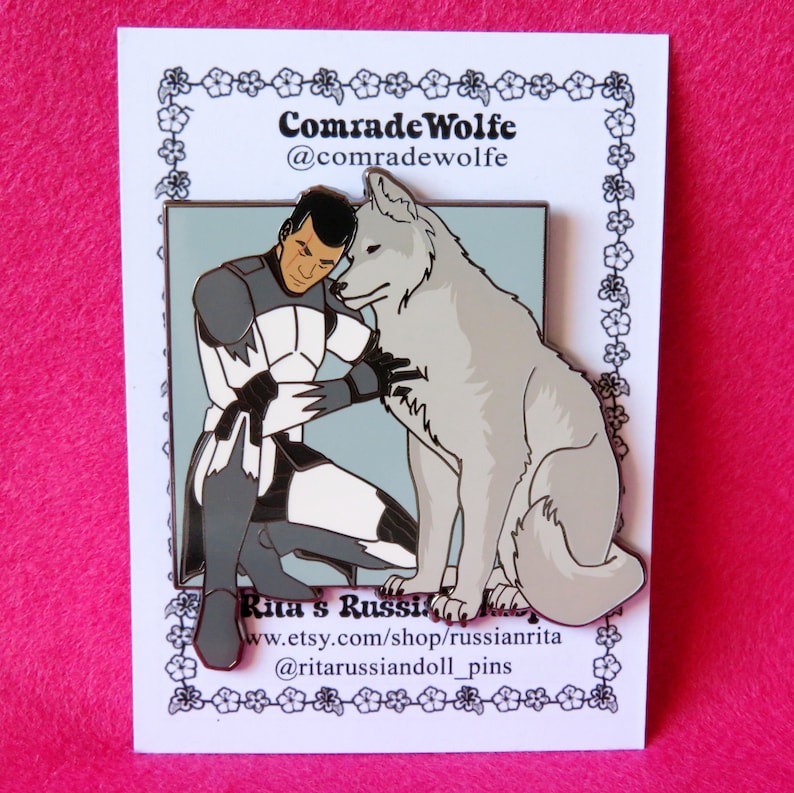 May include: A white enamel pin featuring a cartoon illustration of a man in a gray and white uniform with a white wolf. The man is kneeling and hugging the wolf. The pin has a blue background and the text "Comrade Wolfe @comradewolfe" at the top and "Rita's Russian Army www.etsy.com/shop/russianrita @ritarussiandoll_pins" at the bottom.