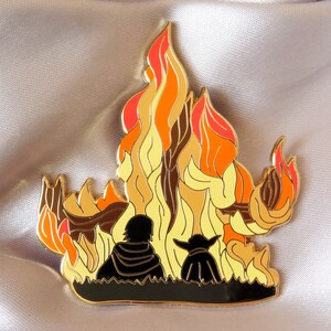 Pu&ograve; includere: Spilla in smalto dorata con due figure in piedi di fronte a un fal&ograve; stilizzato con fiamme arancioni, gialle e rosse.