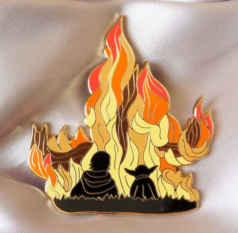 Pu&ograve; includere: Spilla in smalto dorata con due figure in piedi di fronte a un fal&ograve; stilizzato con fiamme arancioni, gialle e rosse.