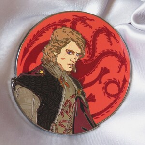 Puede incluir: Un pin de esmalte redondo que presenta un retrato de una persona con cabello largo y rubio, que lleva una chaqueta con estampado marr&oacute;n y dorado y una capa roja. El fondo es un c&iacute;rculo rojo con un dise&ntilde;o de drag&oacute;n de tres cabezas.