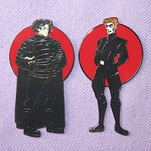 May include: Two enamel pins featuring stylized figures in black attire against red circular backgrounds. One figure has dark hair and a cape, the other has red hair and a white belt. The pins are on a purple background.