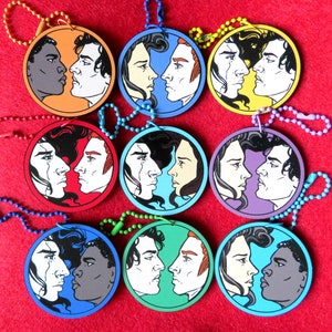 May include: Nine round, colorful keychains with black and white illustrations of couples facing each other. Each keychain features a different color background and a different couple.