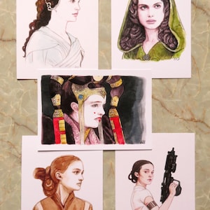 Puede incluir: Conjunto de cinco acuarelas de diferentes personajes de Star Wars. Los personajes son Padmé Amidala, la reina Amidala, Padmé con una capa marrón y Rey Skywalker. Cada pintura está sobre un fondo blanco.