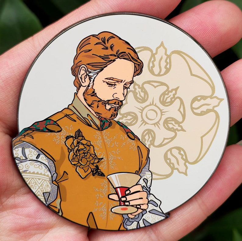 Puede incluir: Un pin de esmalte dorado y blanco con un hombre de pelo rojo y barba que lleva una t&uacute;nica de patr&oacute;n dorado y blanco. Est&aacute; sosteniendo una copa de vino en su mano derecha. El fondo es un c&iacute;rculo blanco con un dise&ntilde;o floral dorado y blanco.