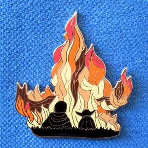 Pu&ograve; includere: Uno spillo in metallo con la silhouette di due figure in piedi di fronte a un fuoco stilizzato arancione, giallo e marrone. Le figure sono rivolte verso l'osservatore.