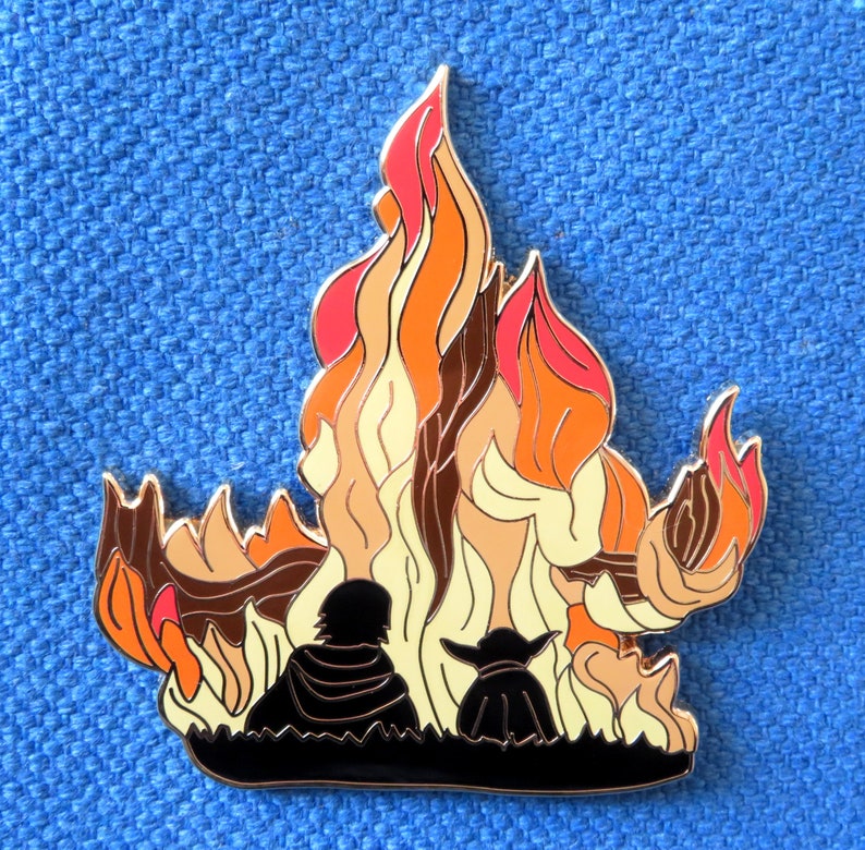 Pu&ograve; includere: Uno spillo in metallo con la silhouette di due figure in piedi di fronte a un fuoco stilizzato arancione, giallo e marrone. Le figure sono rivolte verso l'osservatore.
