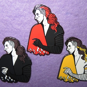 May include: Three enamel pins featuring a stylized portrait of a person with dark hair and a distinctive hairstyle. The pins have a black, red, and yellow color scheme, with the person wearing a black outfit. The pins are set against a purple background.