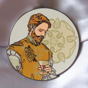Puede incluir: Un pin de esmalte dorado y blanco con un hombre con barba sosteniendo una copa de vino. Lleva una camisa de estampado amarillo y dorado con una flor en el pecho. El fondo es un c&iacute;rculo blanco con un dise&ntilde;o floral dorado.