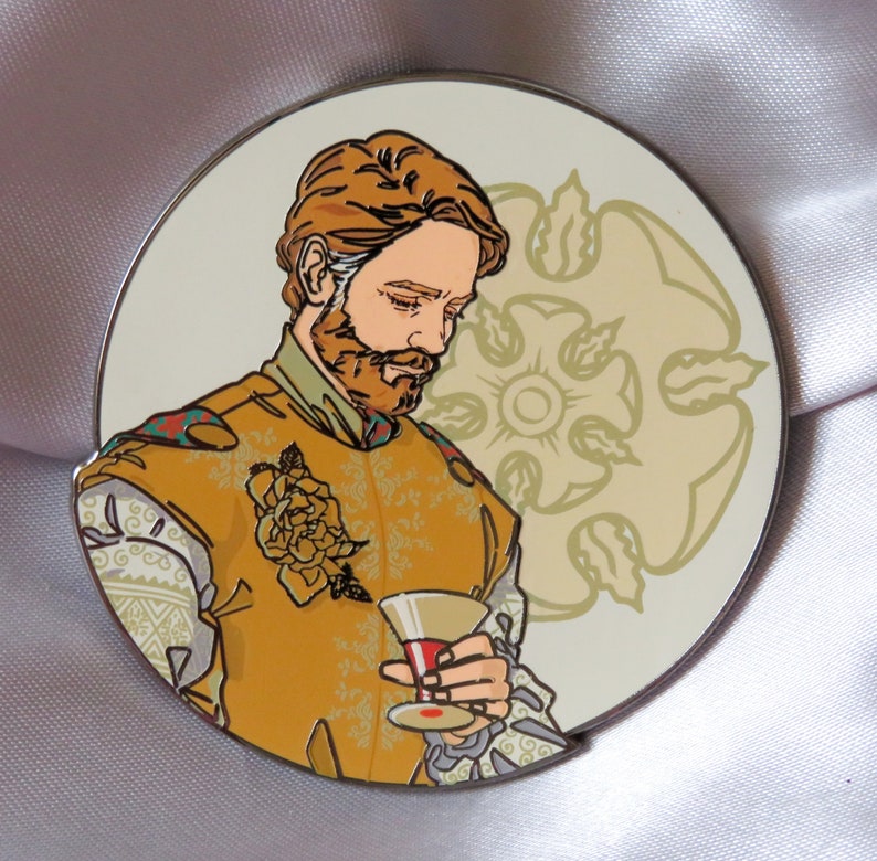 Puede incluir: Un pin de esmalte dorado y blanco con un hombre con barba sosteniendo una copa de vino. Lleva una camisa de estampado amarillo y dorado con una flor en el pecho. El fondo es un c&iacute;rculo blanco con un dise&ntilde;o floral dorado.