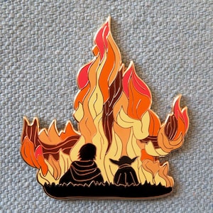 Pu&ograve; includere: Uno spillo in smalto dorato con due figure in piedi di fronte a una fiamma stilizzata arancione e gialla. Le figure sono in silhouette nera.