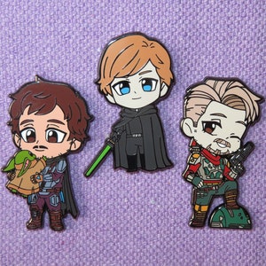 May include: Three enamel pins featuring cartoon characters. One pin depicts a character holding a green baby, another shows a character with a green lightsaber, and the last pin shows a character holding a blaster. The pins are on a purple background.