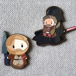 May include: Two enamel pins featuring characters from Star Wars. The first pin is a cartoon of Obi-Wan Kenobi holding a lightsaber. The second pin is a cartoon of Darth Vader holding a lightsaber and carrying a smaller cartoon of a crying Obi-Wan Kenobi.