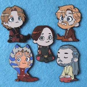 May include: Five enamel pins featuring cartoon characters from the Star Wars franchise. The characters are Obi-Wan Kenobi, Ahsoka Tano, Anakin Skywalker, Yoda, and Qui-Gon Jinn. The pins are all in a chibi style and have a glossy finish.