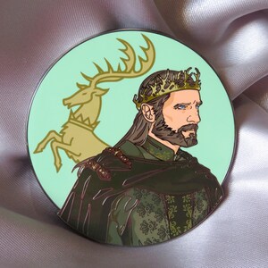 Puede incluir: Un pin de esmalte verde y dorado con el retrato de un hombre con corona y barba. El hombre lleva una t&uacute;nica con estampado verde y dorado. Un ciervo dorado est&aacute; en el fondo.