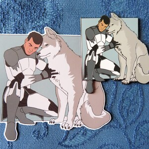 May include: A white wolf with a gray muzzle and a gray tail is sitting next to a man in a white and gray uniform. The man is holding the wolf's neck with his left hand and his right hand is on the wolf's back. The man has short brown hair and a serious expression on his face.