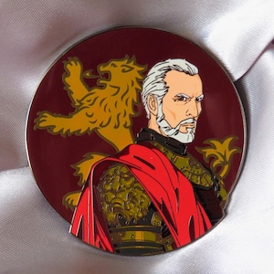 Puede incluir: Un pin de esmalte redondo con el retrato de un hombre con barba blanca y pelo gris. Lleva una capa roja sobre una armadura dorada. Un le&oacute;n dorado est&aacute; en el fondo.