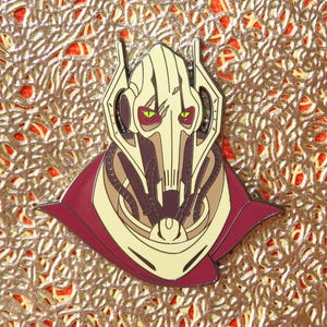 May include: A General Grievous enamel pin featuring a detailed design of the character's head and shoulders. The pin has a cream-colored face with burgundy and brown accents, and green eyes. The pin is set against a textured gold background.