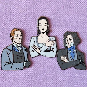 May include: Three enamel pins featuring characters from a classic novel. The pins are black, white, and gray with detailed facial features. The characters are a woman with dark hair, a man with brown hair, and a man with dark hair.