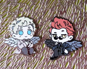 May include: Two enamel pins depicting cartoon characters with angel wings. The character on the left has blonde hair, blue eyes, and is wearing a white suit with a bow tie. The character on the right has red hair, yellow eyes, and is wearing a black suit with a tie and sunglasses.