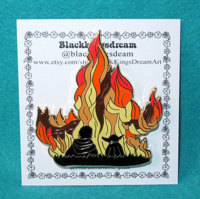 Pu&ograve; includere: Uno spillo in smalto dorato con la silhouette di due figure sedute di fronte a una grande fiamma arancione, gialla e rossa. Lo spillo ha il testo "Black Kings Dream" e "@blackkingsdream" nella parte superiore e "www.etsy.com/shop/BlackKingsDreamArt" nella parte inferiore.