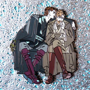 May include: Enamel pin featuring two figures in robes, one with brown hair and the other with auburn hair. The figures are in a seated pose, with one figure's arm around the other. The pin has a silver border and is set against a textured, light blue background.