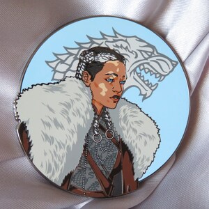 Puede incluir: Un pin de esmalte redondo que presenta a una mujer con un cuello de piel blanca y una cabeza de lobo detr&aacute;s de ella. La mujer tiene el pelo trenzado y lleva una chaqueta de cuero marr&oacute;n con un collar de cadena plateada. La cabeza del lobo es blanca y tiene un fondo azul.