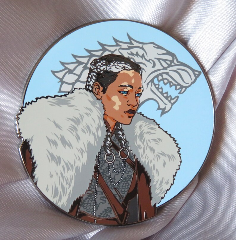 Puede incluir: Un pin de esmalte redondo que presenta a una mujer con un cuello de piel blanca y una cabeza de lobo detr&aacute;s de ella. La mujer tiene el pelo trenzado y lleva una chaqueta de cuero marr&oacute;n con un collar de cadena plateada. La cabeza del lobo es blanca y tiene un fondo azul.