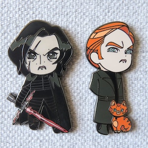 May include: Two enamel pins featuring characters from the Star Wars franchise. The pin on the left is black and depicts Kylo Ren with a red lightsaber. The pin on the right is orange and depicts a character with a ginger beard and a cat.