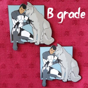 May include: Two enamel pins featuring a white wolf and a man in a white and black uniform. The man is kneeling and petting the wolf. The pins are set against a red fabric background. The text "B grade" is written in white on a black background.