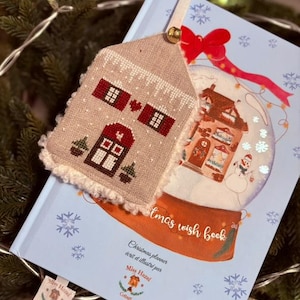 Peut inclure: Un marque-page beige en forme de maison avec des détails rouges et une bordure blanche, posé sur un livre de vœux de Noël bleu clair. Le livre présente une illustration de boule à neige et le texte "Christmas planner écrit et illustré par Miss Hazel". Une petite étiquette est attachée.