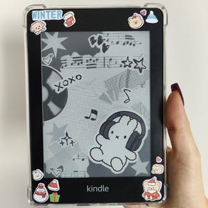 May include: A black Kindle e-reader with a clear protective case. The screen shows a collage of musical notes, stars, and a bunny wearing headphones. Winter-themed stickers are placed around the edges. The word "Kindle" is visible.