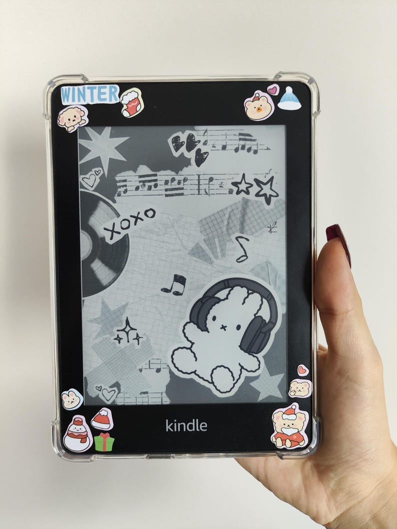 May include: A black Kindle e-reader with a clear protective case. The screen shows a collage of musical notes, stars, and a bunny wearing headphones. Winter-themed stickers are placed around the edges. The word "Kindle" is visible.