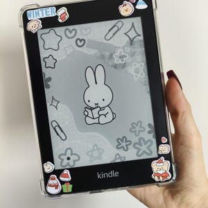 May include: A black Kindle e-reader in a clear protective case, adorned with winter-themed stickers. The screen shows a cartoon bunny reading a book. The word "WINTER" is visible on the top left corner.