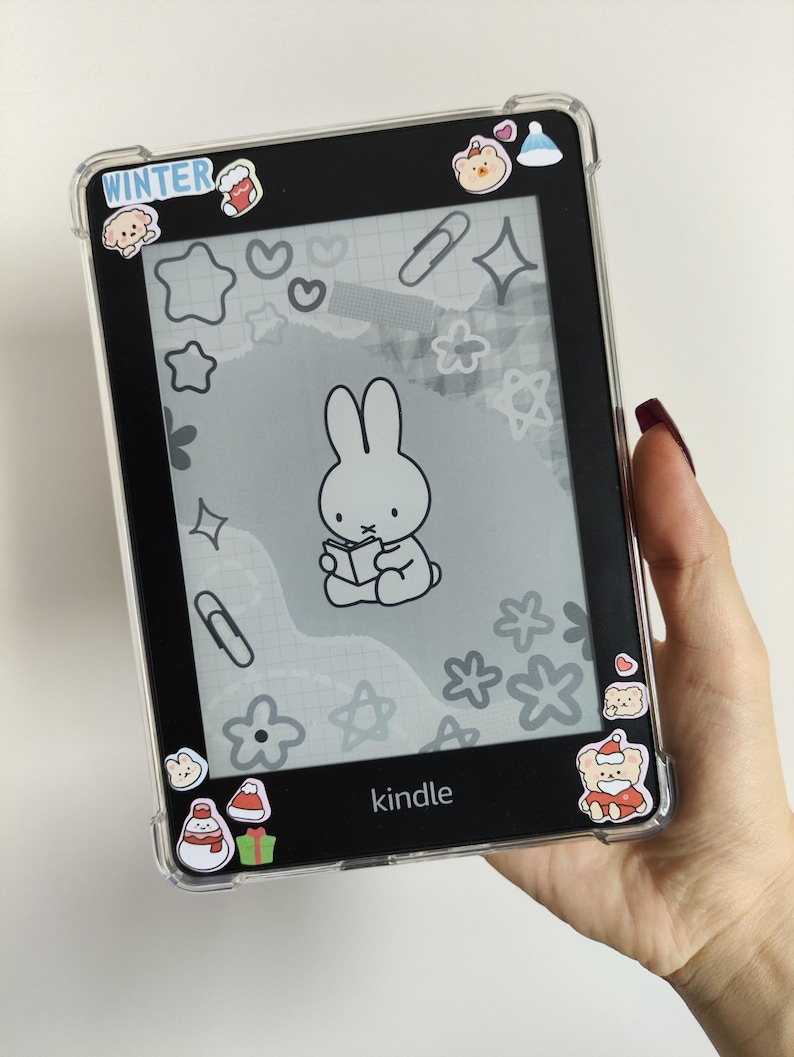 May include: A black Kindle e-reader in a clear protective case, adorned with winter-themed stickers. The screen shows a cartoon bunny reading a book. The word "WINTER" is visible on the top left corner.