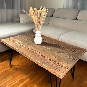 Può includere: Tavolino rettangolare in legno rustico con gambe a forcina nere. Il piano del tavolo presenta un motivo a venature naturali. Un vaso bianco con fiori secchi è sopra. Il divano è di colore chiaro.